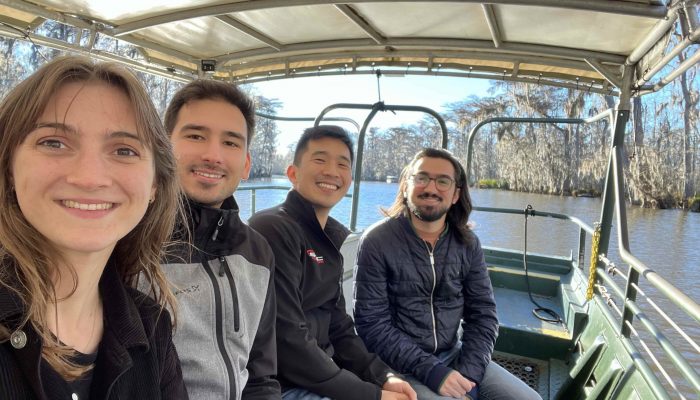4 people sit in a boat and look towards a camera, smiling